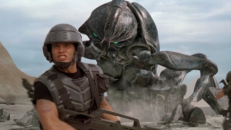 Casper Van Dien as Johnny Rico in Starship Troopers, clad in his military uniform and running away from one of the giant alien bugs that's barreling up behind him.