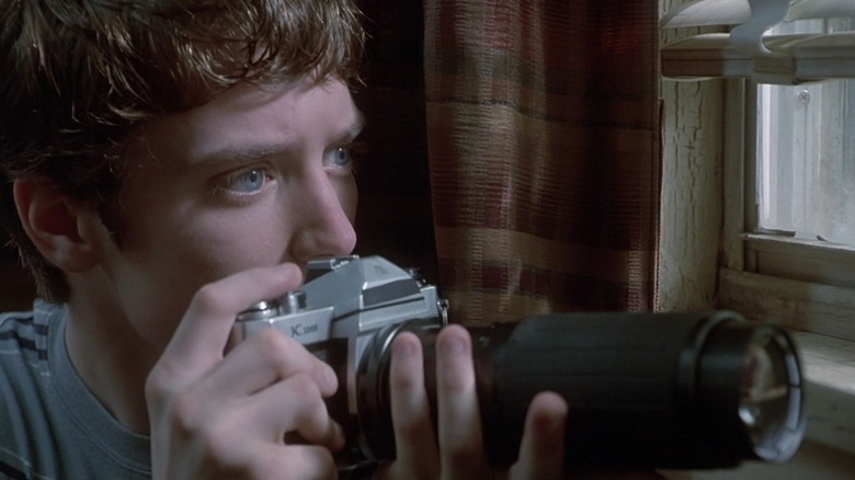 Elijah Wood as Casey Connor in The Faculty, peering through a window while holding a camera with a very long lens up to the lower part of his face.