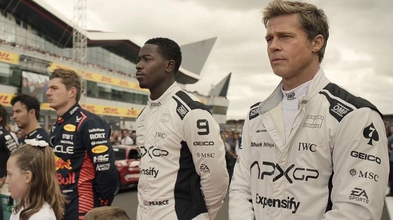 Brad Pitt as Sonny Hayes and Damson Idris as Joshua Pearce in F1, standing side-by-side with other racers in prep for the race.