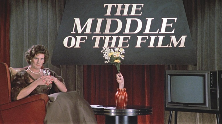 Michael Palin as Female TV Presenter in Monty Python's The Meaning of Life, looking into the camera like a television host on a set with a large sign backdrop that reads "The Middle of the Film."