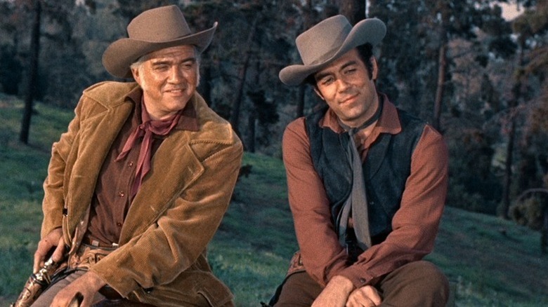 Ben Cartwright (Lorne Greene) and Adam Cartwright (Pernell Roberts) smile while riding on horseback next to each other in Bonanza