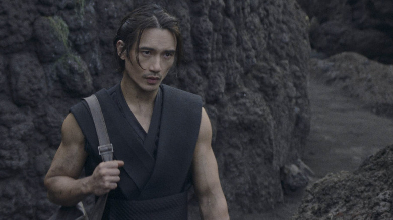 Manny Jacinto as Qimir/The Stranger with his arms exposed standing in front of a large rock on an unknown planet in Star Wars: The Acolyte