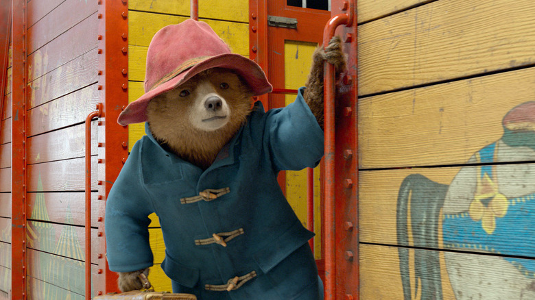 Ben Whishaw as Paddinton in Paddington 2, wearing his coat and red hat as he holds on to a rail on a train.