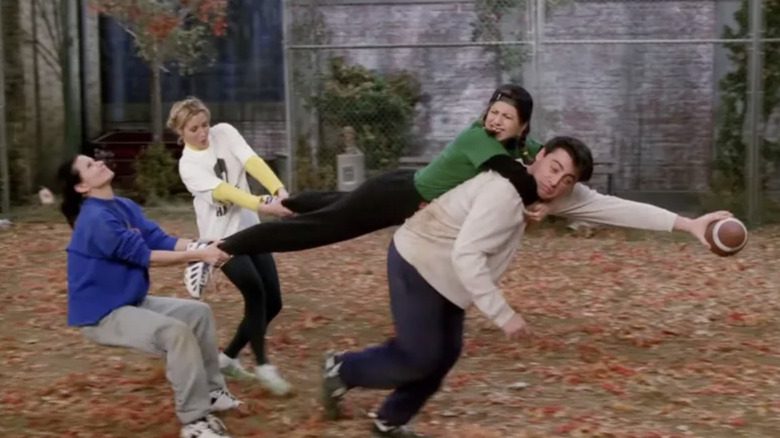 Courteney Cox as Monica Geller, Lisa Kudrow as Phoebe Buffay, and Jennifer Aniston as Rachel Green holding back Joey Tribbiani while playing football in Friends