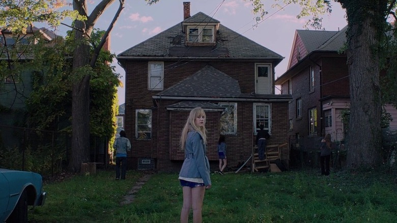 Maika Monroe as Jaime "Jay" Height in It Follows, standing in front of a dilapidated, abandoned house with her friends around it in the background, as she turns and looks behind her toward the camera.