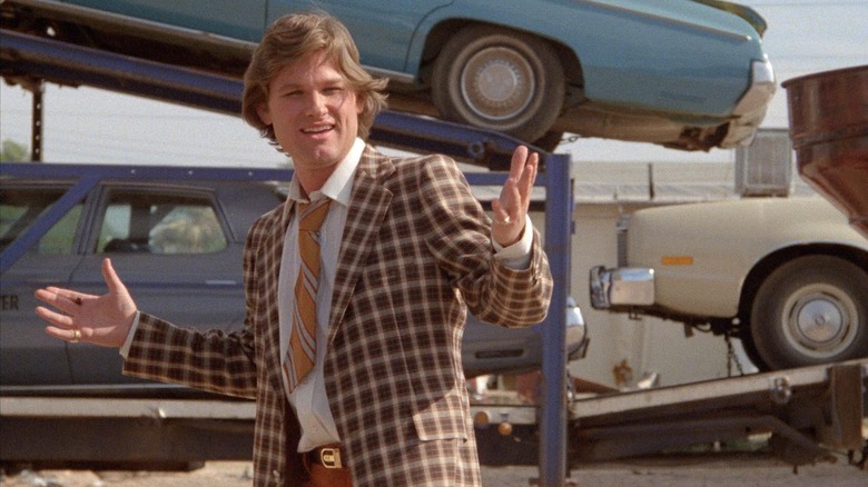 Kurt Russell as Rudy Russo stands in front of a stack of cars in Used Cars