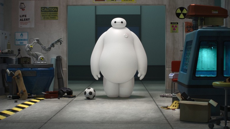 Scott Adsit as Baymax, standing straight up with his arms at his sides in a room with various objects around.