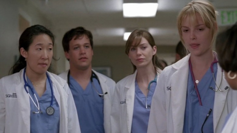 Dr. Cristina Yang (Sandra Oh), Dr, George O'Malley (T.R. Knight), Dr. Meredith Grey (Ellen Pompeo), and Dr. Izzie Stevens (Katherine Heigl) standing in a hospital corridor in Grey's Anatomy