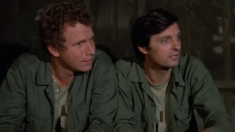 Wayne Rogers as Trapper John McIntyre and Alan Alda as Benjamin Franklin "Hawkeye" Pierce sitting next to each other on M*A*S*H