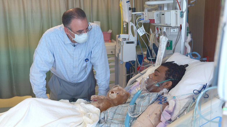 A man wearing a mask examines a man in a bed on a respiratory in Emergency NYC