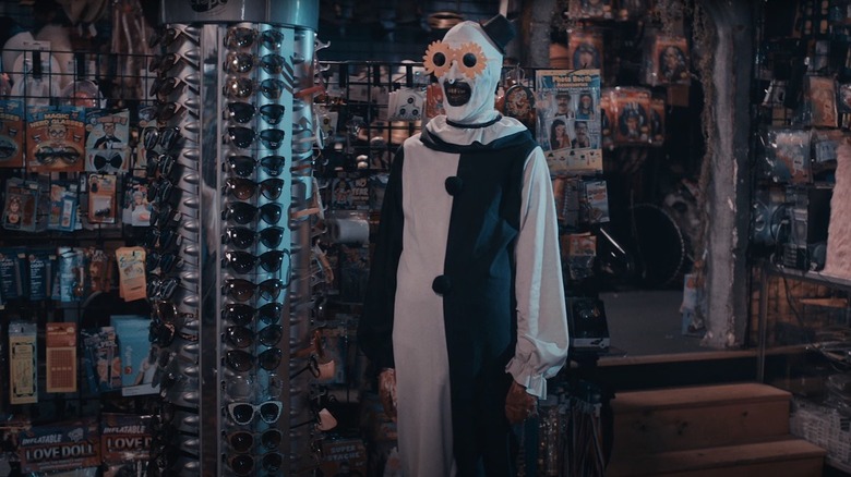 David Howard Thornton as Art the Clown in Terrifier 2, trying on a hip pair of sunflower glasses in a novelty shop.