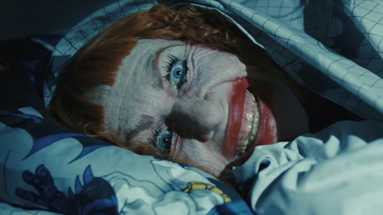 Amy Madigan as Gladys in Weapons, face caked in clownish makeup, smiling and dead-eyed as she lays sideways under the covers in a bed looking right past the camera.