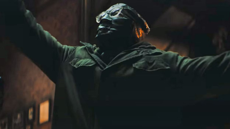 Paul Dano as The Riddler in The Batman, with his trademark jacket and mask on, arms out and looking up toward something.