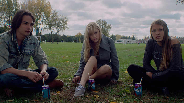 Maika Monroe, Lili Sepe, and Daniel Zovatto in It Follows, sitting next too each other outside in the grass, looking at something out of frame.