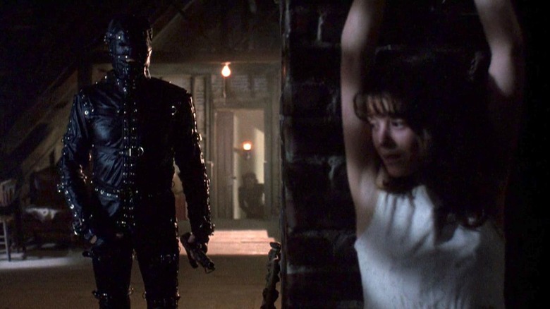 Alice Robeson (A.J. Langer) is bound to a brick column as Eldon Robeson (Everett McGill) approaches in a bondage suit in The People Under the Stairs