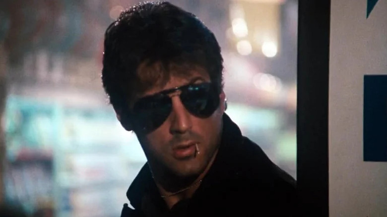 Marion Cobretti (Sylvester Stallone) peers around a corner wearing sunglasses with a matchstick in his mouth in Cobra (1986)