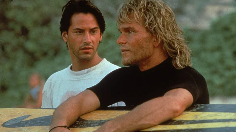 Johnny Utah (Keanu Reeves) looks at Bodhi (Patrick Swayze) as he leans on a surfboard in Point Break (1991)