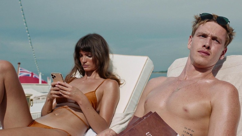 Charlbi Dean as Yaya sitting on pool chair looking at cell phone next to Harris Dickinson as Carl sitting on pool chair holding book in Triangle of Sadness