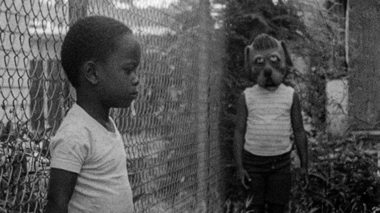 A child stands against a chain-link fence as another child walks up wearing a dog mask in Killer of Sheep