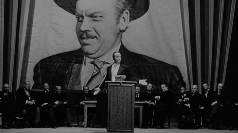 Orson Welles as Charles Foster Kane, standing at a podium with a large group of men behind him, all in front of a large poster of Kane's face.