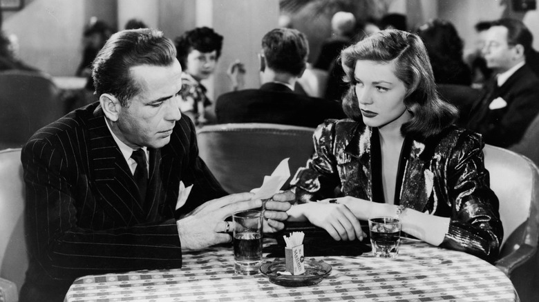 Humphrey Bogart as Philip Marlowe and Lauren Bacall as Vivian Rutledge in The Big Sleep, sitting a table next to each other as Vivian looks at Philip.