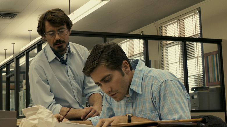 Jake Gyllenhaal as Robert Graysmith and Robert Downey Jr. as Paul Avery in Zodiac, sitting in an office together while Robert looks down at something on his desk and Paul looks on at him.