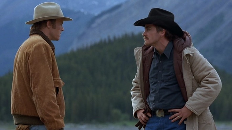 Heath Ledger as Ennis Del Mar and Jake Gyllenhaal as Jack Twist in Brokeback Mountain, both in their cowboy gear against the forested mountains, looking at each other with some frustration, as Jack puts his hands on his hips.