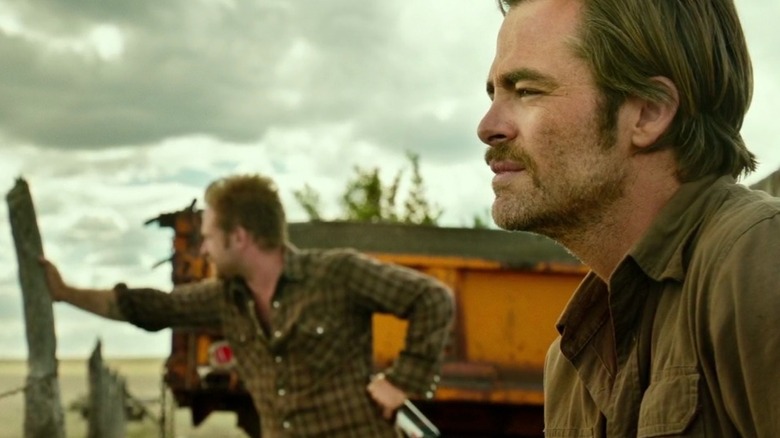 A profile shot of Chris Pine as Toby in the foreground and Ben Foster as Tanner in the background, both standing outside by a barbed wire fence and looking out.