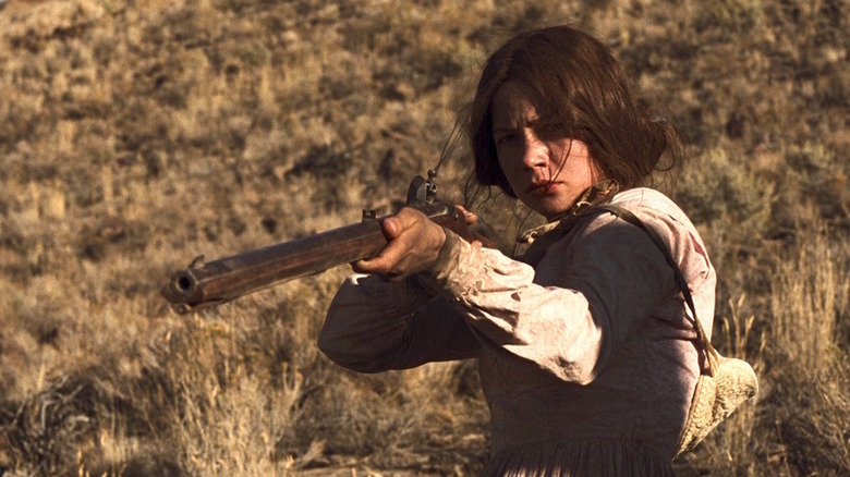 Michelle Williams as Emily Tetherow in Meek's Cutoff, standing in an arid plaid and pointing a rifle off at someone out of frame.