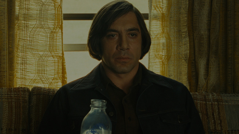 Javier Bardem as Anton Chigurh in No Country For Old Men, sitting on a couch in front of a window.
