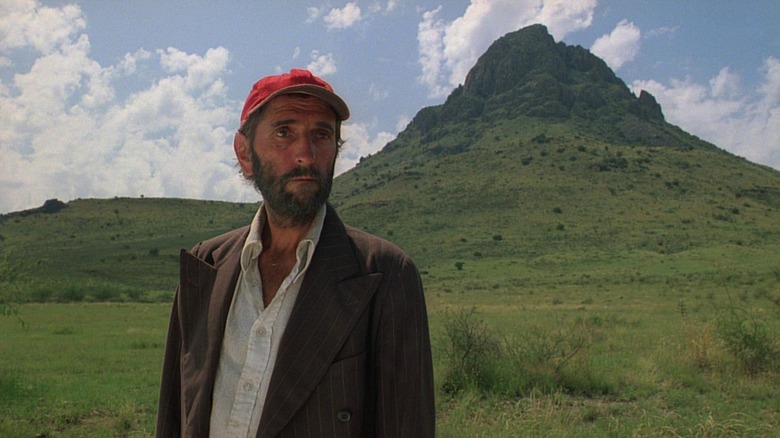 Harry Dean Stanton as Travis Henderson in Paris, Texas, looking dirty and disheveled in his shirt, jacket, and ball cap as he stands outside against a grassy mountain.