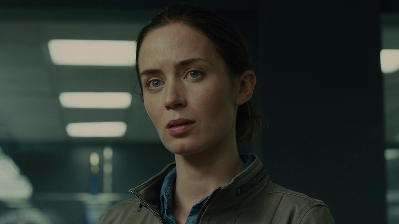 Emily Blunt as Kate Macer in Sicario, standing in a non descript room and looking on at something.