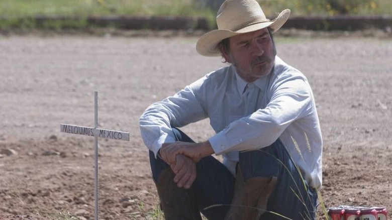 Tommy Lee Jones as Pete Perkins in The Three Burials of Melquiades Estrada, sitting with his button-up Western shirt and large-brimmed hat on, sitting next to a makeshift cross in the ground that says Melquiades Estrada on it.