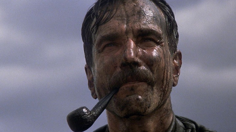 A close-up of Daniel Day-Lewis as Daniel Plainview in There Will Be Blood, framed against an overcast sky with a pipe in his mouth and covered in oil as he looks on.