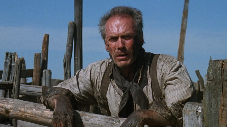 Clint Eastwood as William "Will" Munny in Unforgiven, standing with the sun shining in his face as he squints his eyes and leads over a wood post railing with dirtied hands.