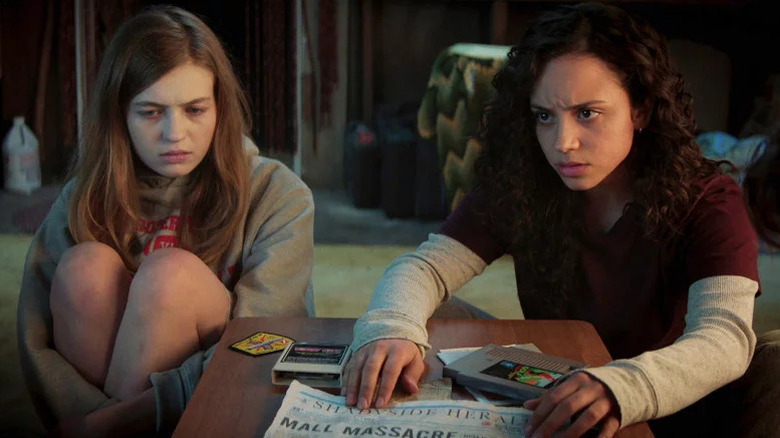 Olivia Scott Welch as Sam in grey sweatshirt with legs at chest next to Kiana Madeira as Deena in long-sleeve shirt holding newspaper in Fear Street Part One: 1994