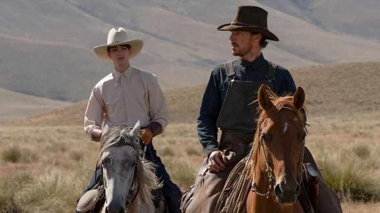 Kodi Smit-McPhee as Peter in white shirt and hat on white horse next to Benedict Cumberbatch as Phil in dark hat and dark clothings on brown horse in The Power of the Dog