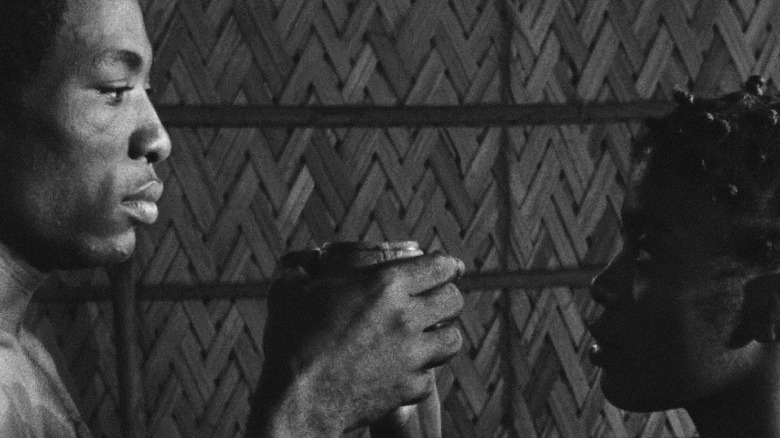David Endene as Ngando and Arlette Din Bell as Ndomé locking eyes while holding the same cup in Muna Moto
