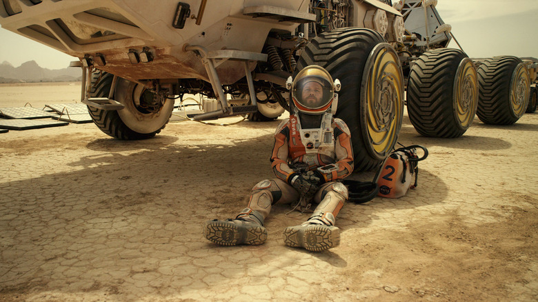 Matt Damon as Dr. Mark Watney, lying against the wheel of a mars vehicle in The Martian.