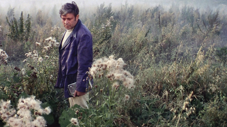 Donatas Banionis as Kris Kelvin  weaing a jacket and holding a device while standing in a field in Solaris