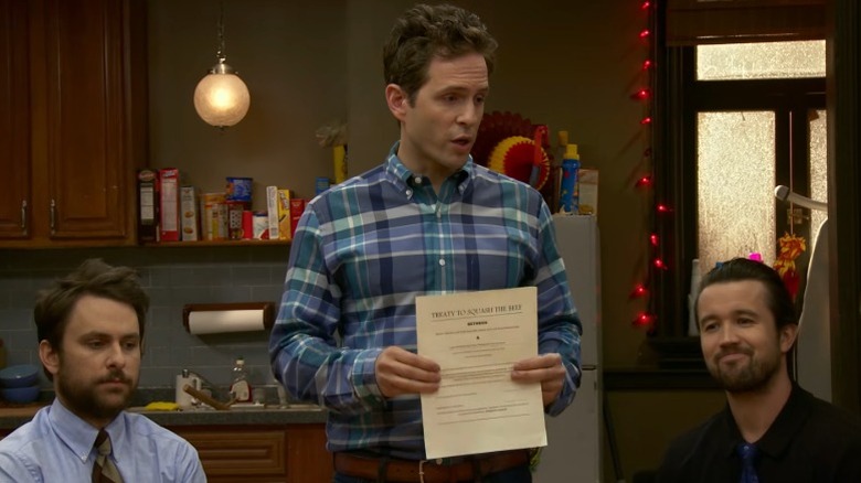 Glenn Howerton as Dennis Reynolds standing between a sitting Charlie Day as Charlie Kelly and Rob McElhenney as Mac and holding a document It's Always Sunny in Philadelphia
