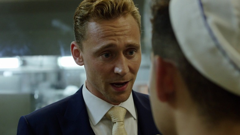 Tom Hiddleston as Jonathan Pine wearing a blue suit and talking to a hotel chef