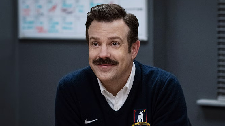 Jason Sudeikis as Ted Lasso, smiling in the managers office on Ted Lasso