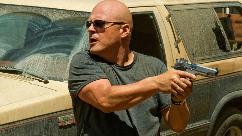 Michael Chiklis as Vic Mackey, holding a gun and taking cover behind a car in The Shield.