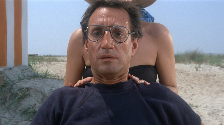 Roy Scheider as Martin Brody in Jaws, sitting on the beach with as a woman rubs his shoulders and he looks on in concern.