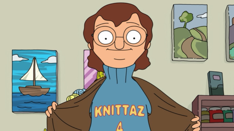 A man in glasses holds open his jacket, displaying a sweater that reads "Knittaz 4 Life"
