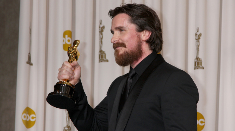 Christian Bale in black suit holding up Oscar with Oscar statue behind him in 2011