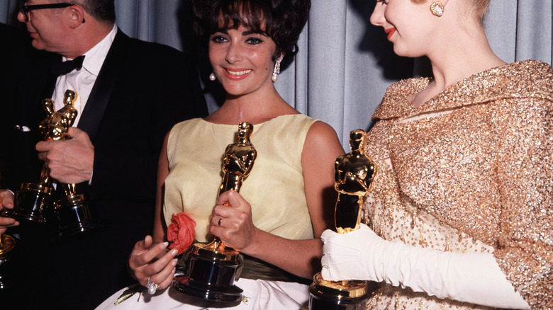 Burt Lancaster looking away in tux holding two Oscars next to Elizabeth Taylor in yellow dress holding Oscar and Shirley Jones in glittery gold dress holding Oscar in 1961