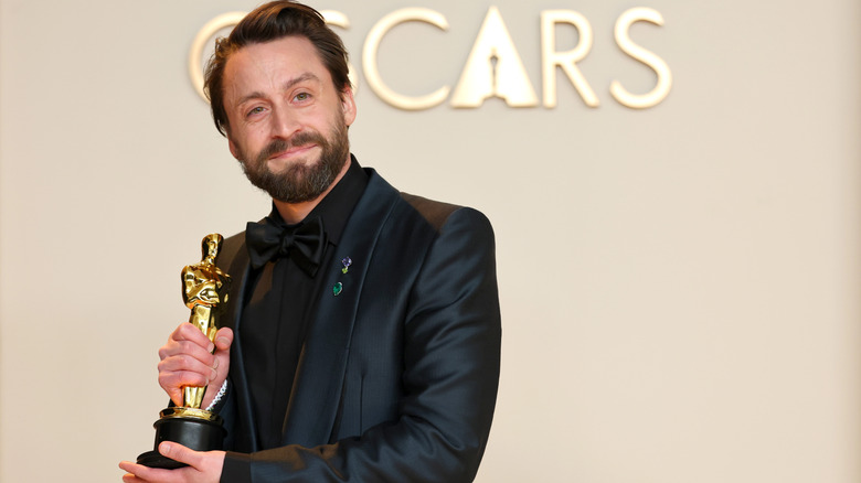 Kieran Culkin in black suit smiling and holding Oscar in front of beige background in 2025