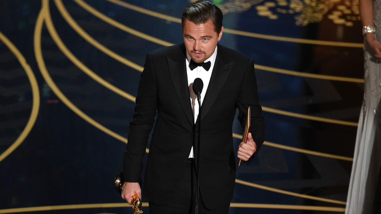 Leonardo DiCaprio on stage standing in front of microphone with Oscar in hand wearing tux in 2016